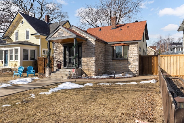 snow covered back of property featuring stone siding, a shingled roof, a chimney, and fence