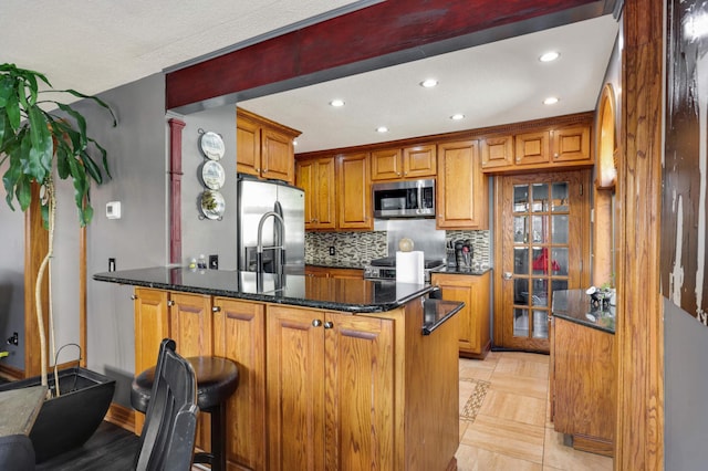 kitchen featuring dark stone countertops, a peninsula, appliances with stainless steel finishes, brown cabinets, and backsplash