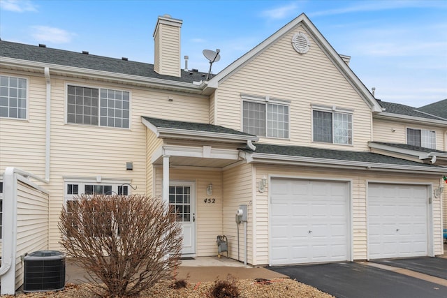 view of front of property featuring a chimney, a shingled roof, an attached garage, central AC unit, and driveway