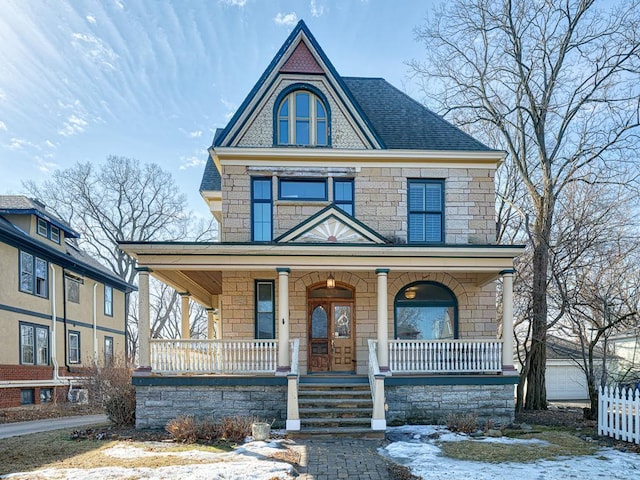 victorian-style house with stone siding, roof with shingles, a porch, and fence