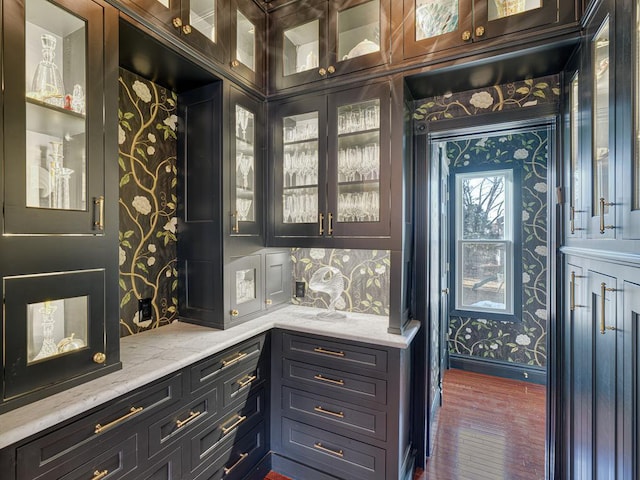 kitchen with light stone counters, baseboards, wallpapered walls, dark wood-style flooring, and glass insert cabinets