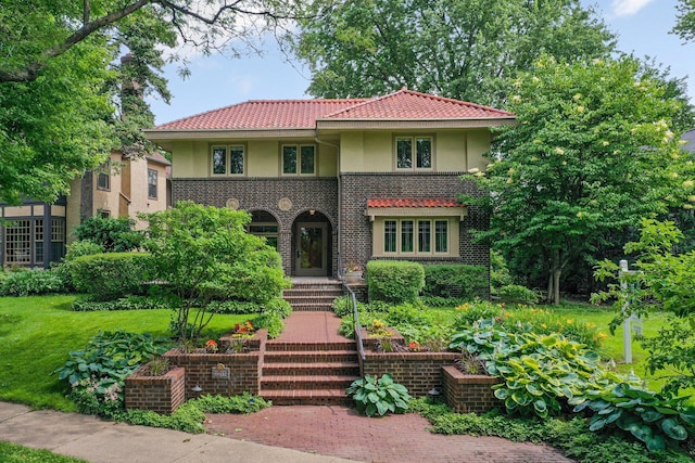 mediterranean / spanish-style home featuring a tiled roof, brick siding, a front yard, and stucco siding