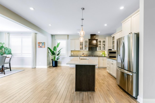 kitchen with stainless steel appliances, custom range hood, decorative backsplash, an island with sink, and light wood-type flooring