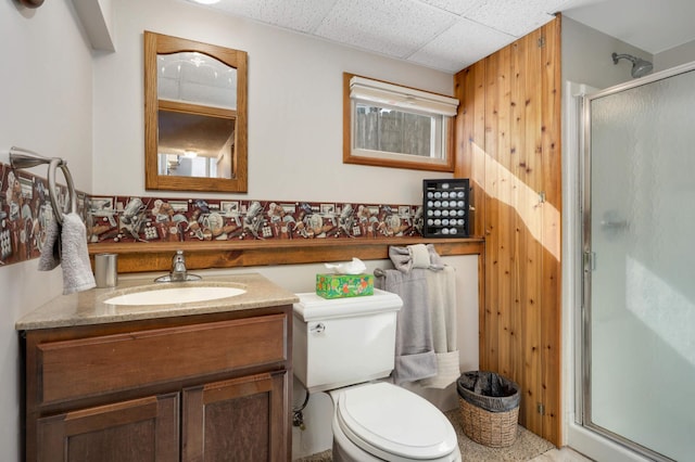 bathroom featuring toilet, a stall shower, wooden walls, vanity, and a drop ceiling