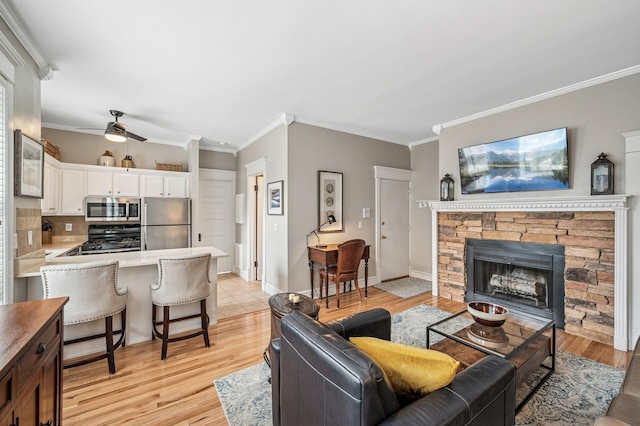 living room featuring a ceiling fan, baseboards, light wood finished floors, ornamental molding, and a stone fireplace