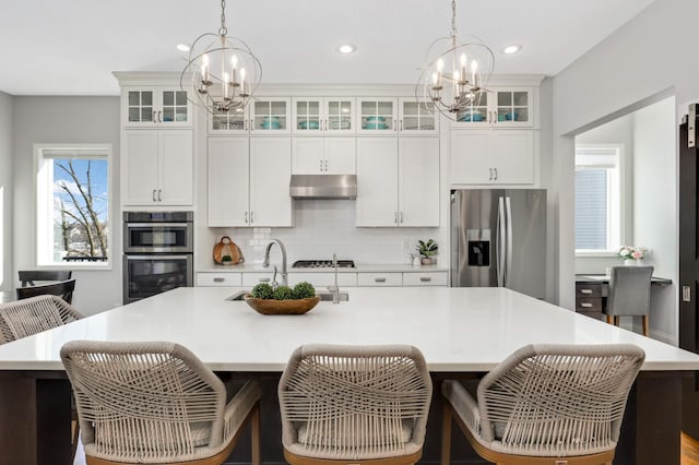 kitchen featuring an island with sink, appliances with stainless steel finishes, a notable chandelier, under cabinet range hood, and a sink