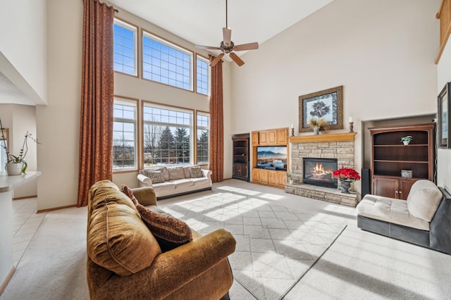 living area with baseboards, ceiling fan, a stone fireplace, a towering ceiling, and light colored carpet