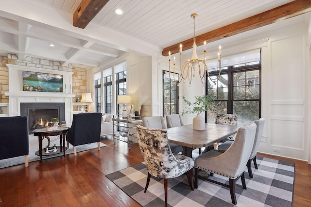 dining space with wood finished floors, a warm lit fireplace, beamed ceiling, a decorative wall, and a chandelier