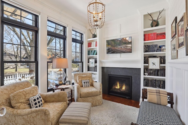 sitting room with built in features, a lit fireplace, ornamental molding, an inviting chandelier, and wood finished floors