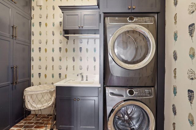 laundry room with stacked washing maching and dryer, wallpapered walls, cabinet space, brick floor, and a sink