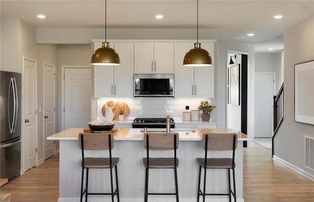 kitchen with light wood finished floors, decorative backsplash, a kitchen island with sink, stainless steel appliances, and light countertops