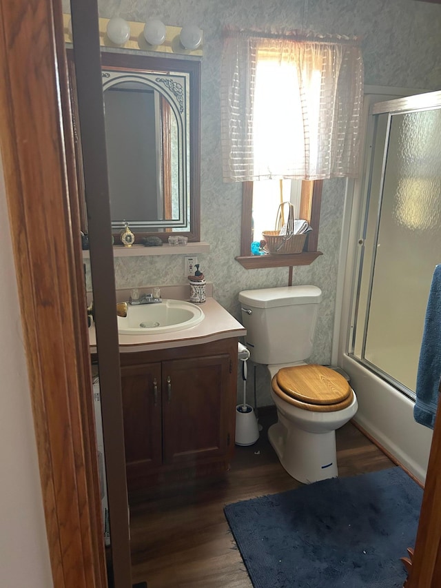 bathroom featuring vanity, wood finished floors, wallpapered walls, toilet, and combined bath / shower with glass door