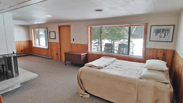 carpeted bedroom with a wainscoted wall, wood walls, a multi sided fireplace, visible vents, and baseboard heating