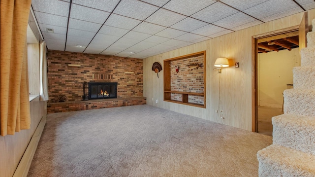 unfurnished living room with wooden walls, a baseboard radiator, a fireplace, a drop ceiling, and carpet flooring