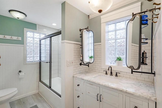 bathroom featuring vanity, wood finished floors, shower / bath combination with glass door, wainscoting, and toilet