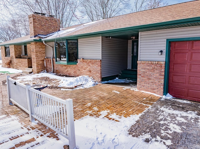 snow covered property entrance with brick siding, a chimney, a garage, and a shingled roof