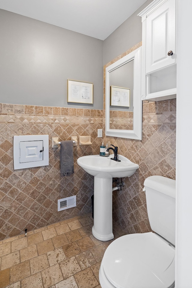 half bath with visible vents, toilet, stone tile floors, tile walls, and a sink