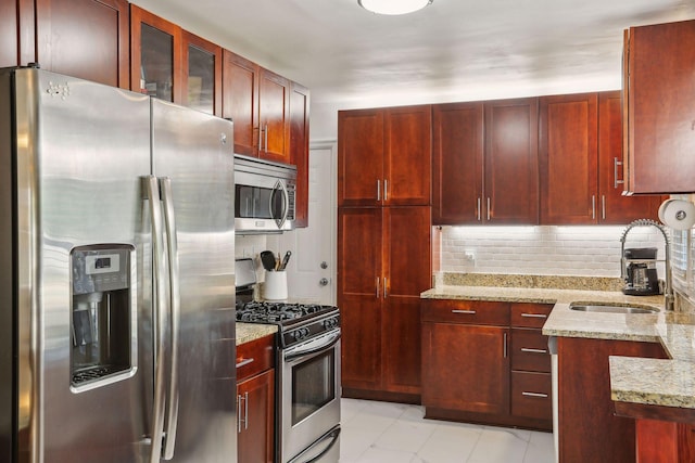 kitchen with light stone counters, a sink, decorative backsplash, appliances with stainless steel finishes, and reddish brown cabinets