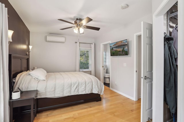 bedroom featuring a wall unit AC, baseboards, ceiling fan, ensuite bathroom, and light wood-type flooring