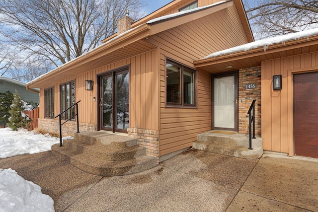 snow covered property entrance with brick siding, stone siding, board and batten siding, and a chimney