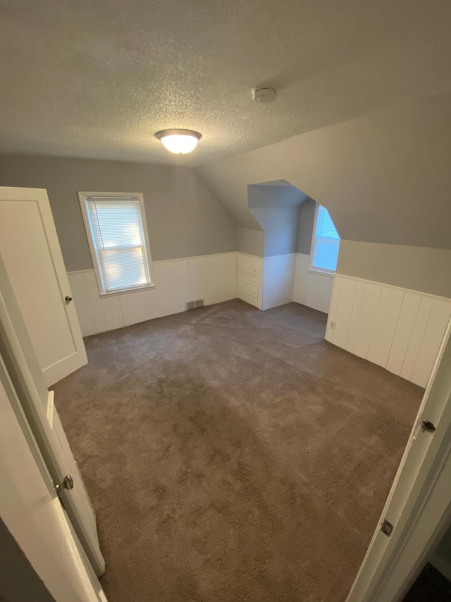 bonus room featuring visible vents, lofted ceiling, wainscoting, a textured ceiling, and dark colored carpet