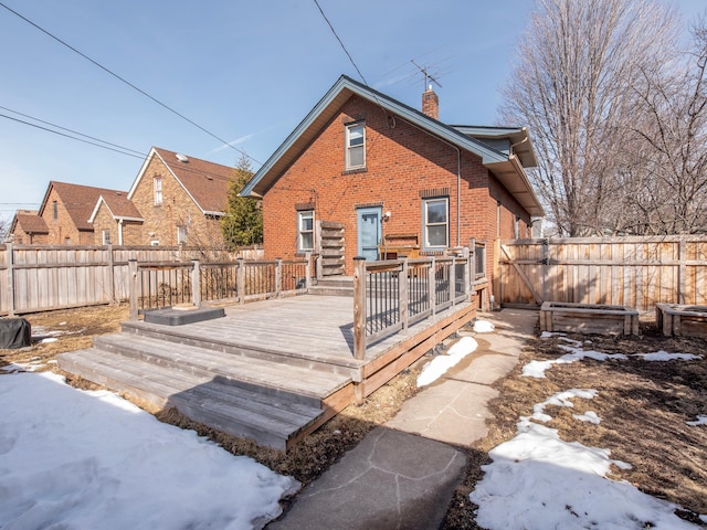 snow covered back of property featuring a wooden deck, a fenced backyard, brick siding, and a chimney