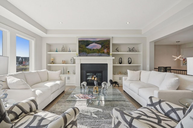 living room with a glass covered fireplace, a tray ceiling, built in shelves, and wood finished floors