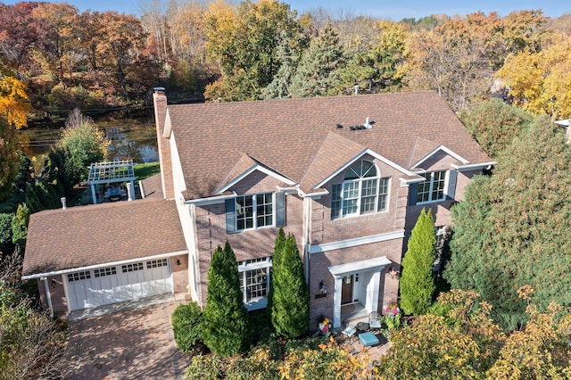 view of front of house featuring driveway, a view of trees, a shingled roof, a garage, and a chimney