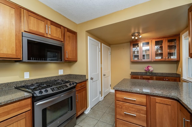 kitchen with brown cabinets, a textured ceiling, stainless steel appliances, light tile patterned flooring, and glass insert cabinets