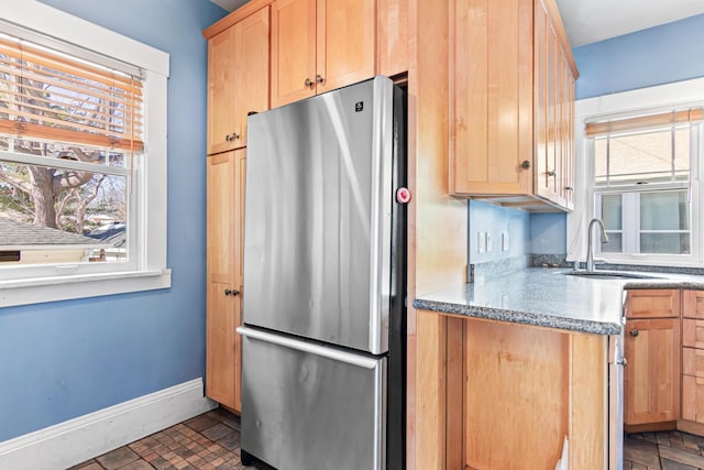 kitchen with light brown cabinetry, a sink, freestanding refrigerator, brick floor, and baseboards