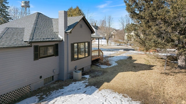 view of snowy exterior with central air condition unit, a chimney, and a shingled roof