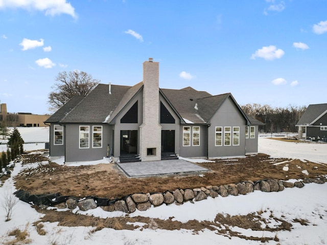 snow covered rear of property featuring entry steps, roof with shingles, and a chimney