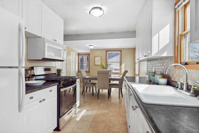 kitchen with dark countertops, backsplash, white cabinets, white appliances, and a sink