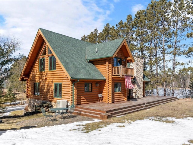 snow covered rear of property with log siding, central air condition unit, a shingled roof, and a balcony