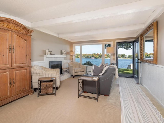 living area featuring a water view, beam ceiling, a fireplace with raised hearth, wainscoting, and light colored carpet