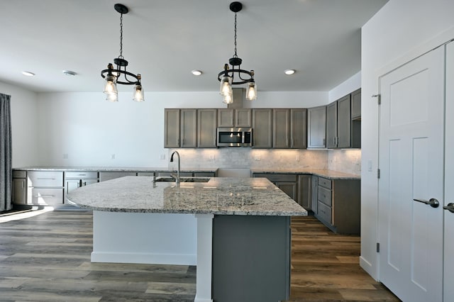 kitchen with stainless steel microwave, backsplash, dark wood-type flooring, a center island with sink, and a sink