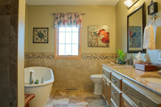 bathroom with toilet, stone tile floors, tile walls, a freestanding bath, and vanity