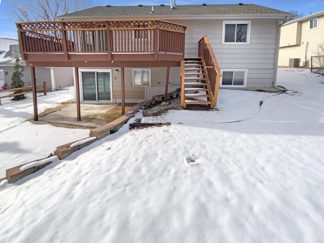 snow covered house with a deck, cooling unit, a shingled roof, stairs, and a patio area