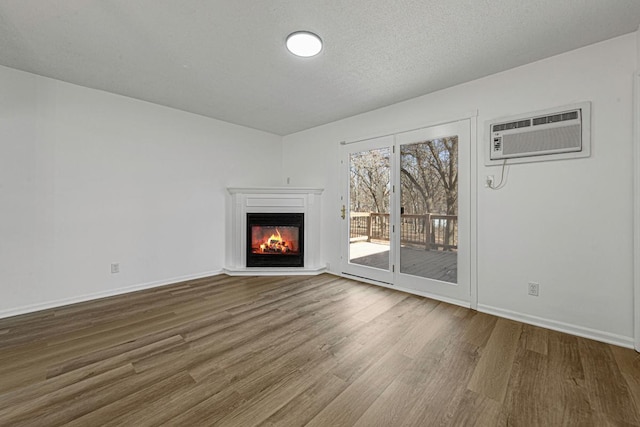 unfurnished living room featuring baseboards, wood finished floors, a glass covered fireplace, a textured ceiling, and a wall mounted AC