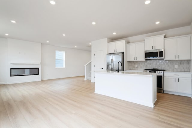 kitchen with backsplash, an island with sink, stainless steel appliances, a glass covered fireplace, and white cabinetry