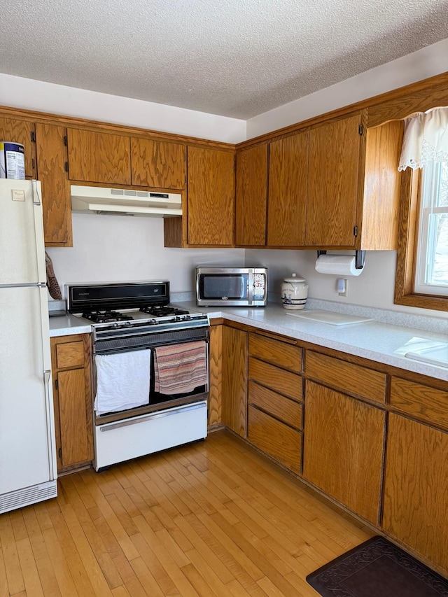 kitchen featuring stainless steel microwave, under cabinet range hood, freestanding refrigerator, brown cabinetry, and gas stove