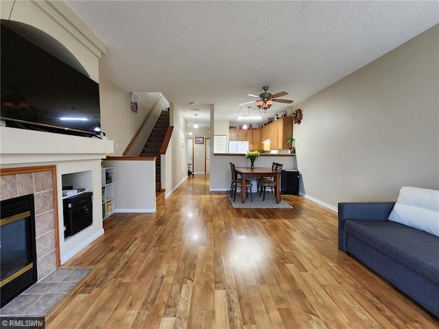 living room with stairway, light wood-style flooring, a tile fireplace, a textured ceiling, and a ceiling fan