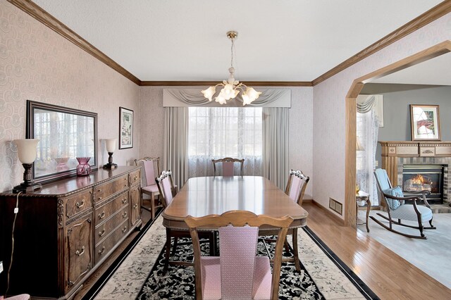 dining space featuring light wood finished floors, wallpapered walls, an inviting chandelier, ornamental molding, and a glass covered fireplace