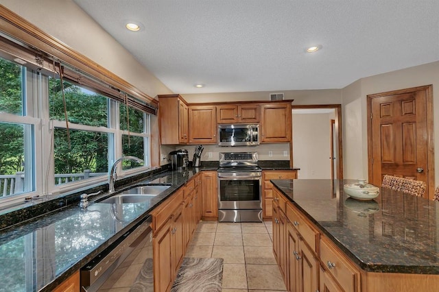 kitchen featuring light tile patterned floors, dark stone countertops, brown cabinets, stainless steel appliances, and a sink