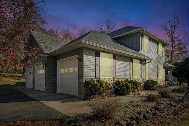 property exterior at dusk with driveway, a garage, stone siding, and roof with shingles