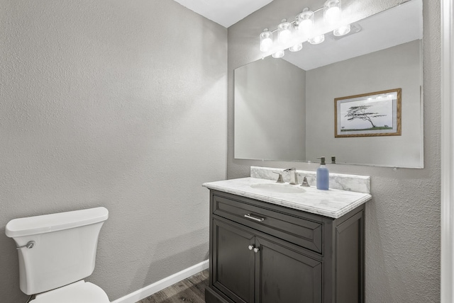 half bathroom with vanity, wood finished floors, baseboards, toilet, and a textured wall