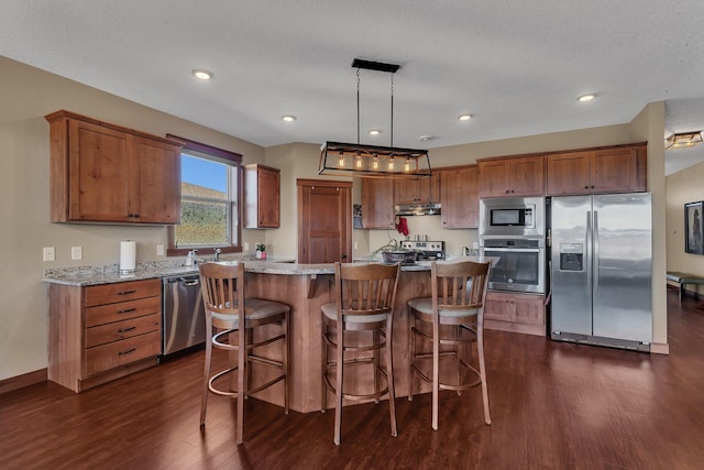 kitchen with brown cabinetry, dark wood-type flooring, under cabinet range hood, appliances with stainless steel finishes, and a center island