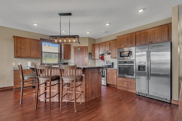 kitchen with brown cabinetry, under cabinet range hood, a kitchen bar, appliances with stainless steel finishes, and dark wood-style floors