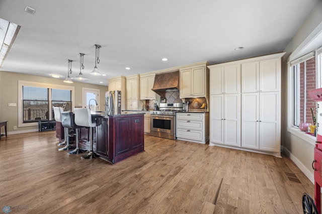 kitchen with light wood-style flooring, an island with sink, custom range hood, backsplash, and stainless steel appliances