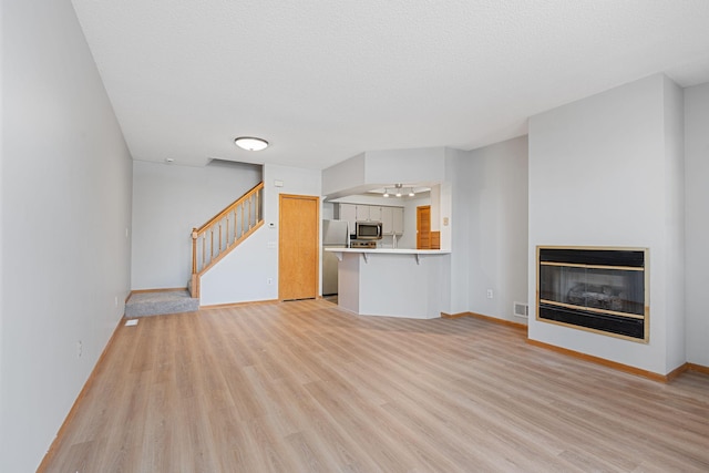 unfurnished living room featuring light wood-type flooring, visible vents, a glass covered fireplace, stairway, and baseboards